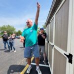 Smiling man in a teal shirt raises a small object above his head during an outdoor gathering in a parking lot while others watch.