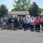Large group of people standing together outdoors in a parking lot, posing for a group photo in front of a small building on a sunny day with trees in the background.
