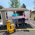 Two people stand outside a small shed, holding a large CRUTCHMAN sign with yellow DeWalt tool boxes on the pavement nearby.