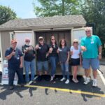 Seven volunteers stand in front of an open trailer, giving thumbs up at an outdoor event.