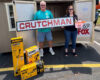 Two volunteers hold a large CRUTCHMAN sign in front of a trailer, with DeWalt tool boxes on the pavement and a 69 The Fox banner nearby.