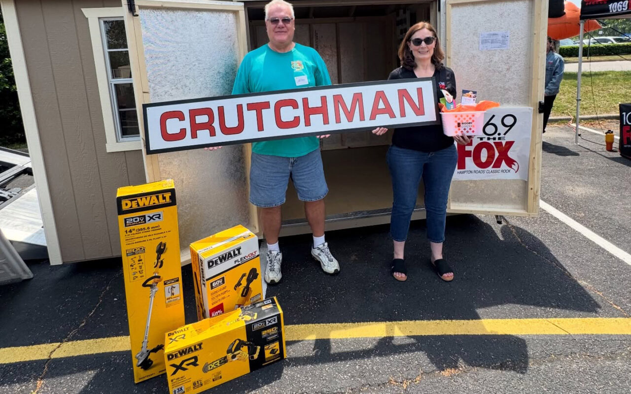 Two volunteers hold a large CRUTCHMAN sign in front of a trailer, with DeWalt tool boxes on the pavement and a 69 The Fox banner nearby.
