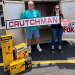 Two volunteers hold a large CRUTCHMAN sign in front of a trailer, with DeWalt tool boxes on the pavement and a 69 The Fox banner nearby.