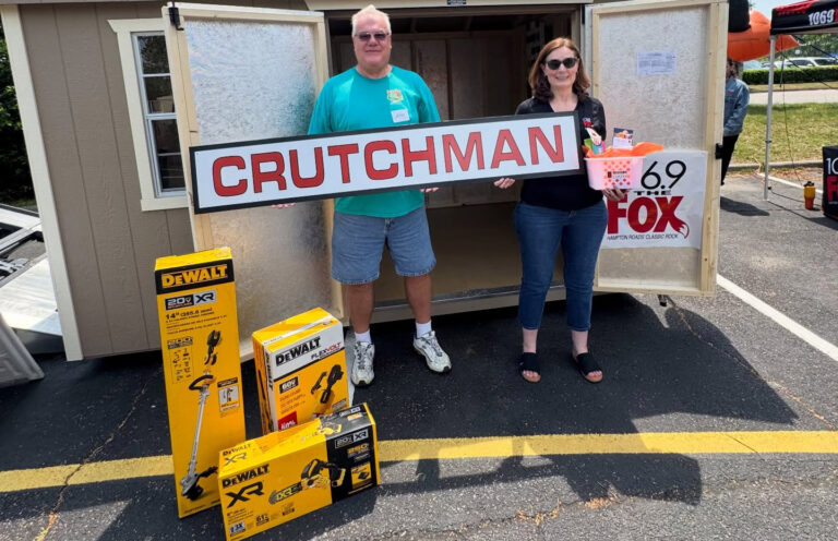 Two volunteers hold a large CRUTCHMAN sign in front of a trailer, with DeWalt tool boxes on the pavement and a 69 The Fox banner nearby.